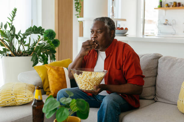 Watching television and eating popcorn. African pensioner sitting at his cozy home, watching television and eating popcorn. He is enjoying the afternoon. streaming service stock pictures, royalty-free photos & images