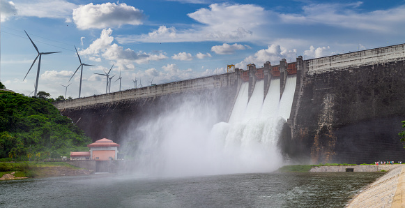 Water over spillway of the concrete dam and have wind turbines with blue sky on top , both process are working for renewable and clean energy.
