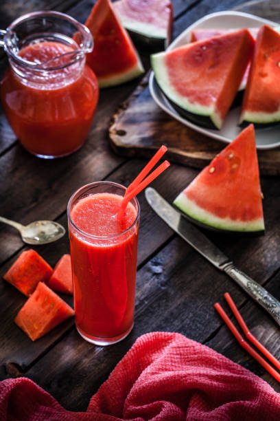 Watermelon juice with slices on rustic wooden table High angle view of a tall glass with two red drinking straws filled with watermelon juice shot on rustic wooden table. Some watermelon slices are in a white plate and placed all directly on the table around the glass. Predominant colors are red and brown. Low key DSRL studio photo taken with Canon EOS 5D Mk II and Canon EF 100mm f/2.8L Macro IS USM. watermelon juice stock pictures, royalty-free photos & images