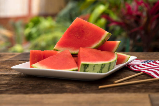 Watermelon triangle sections for healthy summertime snack This is an outdoor photograph of sliced red see this watermelon on a white square modern plate sitting on a wooden picnic bench outdoors for a simple and concept of healthy eating and snacks during the summertime fun. memorial day background stock pictures, royalty-free photos & images