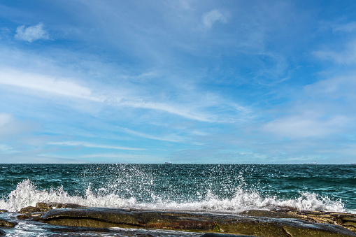 Waves crashing against flat volcanic rock with ships on the horizon underneath pretty blue sky with fluffy clouds.