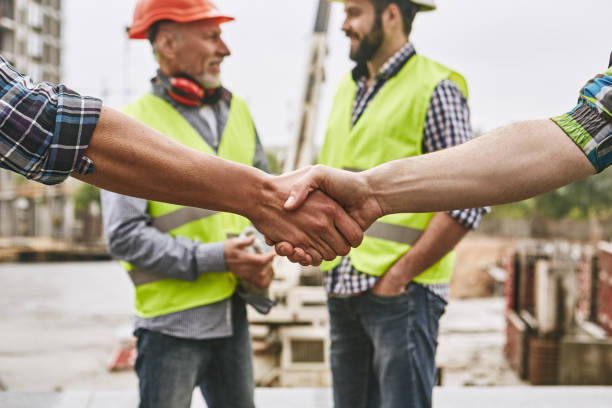 We did it! Close up photo of builders shaking hands against cheerful colleagues while working together at construction site. Team work. stock photo
