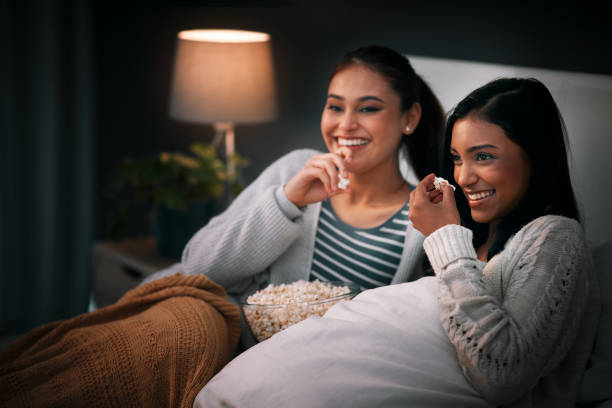 We never watch an episode alone Shot of two young woman eating popcorn while sitting together on a bed at night asian family eating together stock pictures, royalty-free photos & images