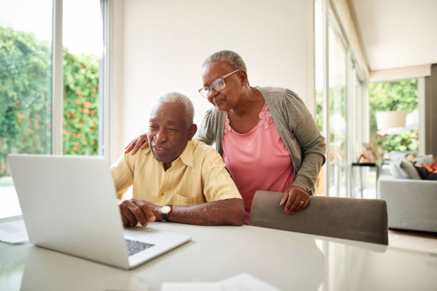 We understand the importance of investing money Shot of african senior man working on laptop with his wife standing by at home laptop couple stock pictures, royalty-free photos & images
