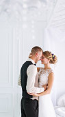Wedding couple in love. Beautiful bride in white dress and handsome groom suite standing and embracing each other indoors in decorated studio room, white bright interior