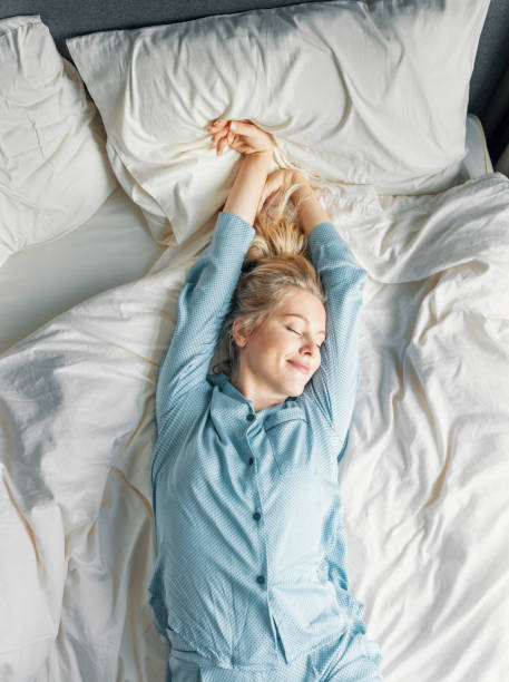 Weekend Mornings: a Beautiful Blonde Woman in Blue Pajamas Lying in Bed in the Morning Waking up: a beautiful happy woman stretches in bed in the morning after a good night's sleep, eyes closed, an overhead view. sleep with long hair stock pictures, royalty-free photos & images
