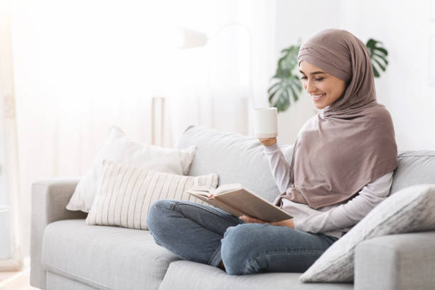 Weekend Pastime. Young Arab Woman Relaxing With Book And Coffee On Couch Weekend Pastime. Young Arab Woman In Hijab Relaxing With Book And Coffee On Couch, Enjoying Spending Time At Home, Copy Space hot arab women stock pictures, royalty-free photos & images