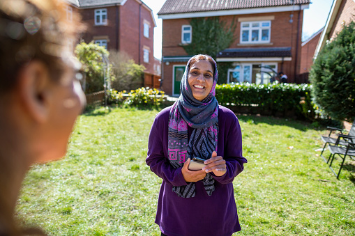 Over the shoulder view of two female friends standing in a back garden owned by one of them, talking face to face with one another. One woman is wearing a headscarf.