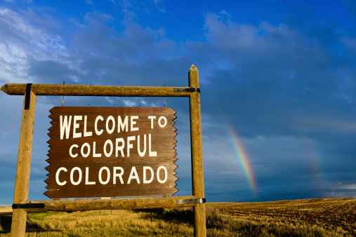 A wooden sign on the border of Colorado welcoming people into the state.  There is a double rainbow in the background over fields
