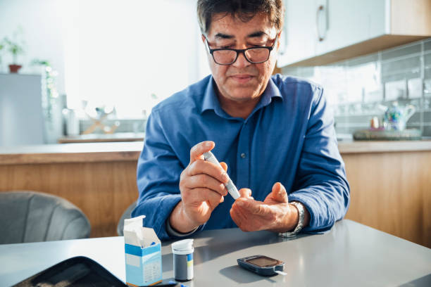 Wellness and Healthcare A mature Asian man sitting at a dining table in his kitchen, he is pricking his finger using a glaucometer to test his blood sugar levels, he is managing his diabetes. diabetes stock pictures, royalty-free photos & images
