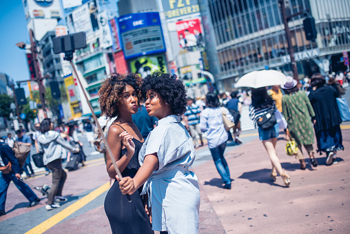 Two young women taking selfie on a city street in Shibuya crossing, Tokyo, Japan. They are sending kisses, looking at mobile phone and using selfie stick.