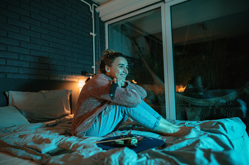 Photo of a smiling young woman while watching TV in the bedroom of her apartment; eating sushi and enjoying her night at home alone.