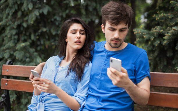 What is in your phone concept Young couple in the park. Boy is checking his phone hiding it. Girl is lookin into boyfriend's phone with mistrust. What is in your phone concept envy stock pictures, royalty-free photos & images