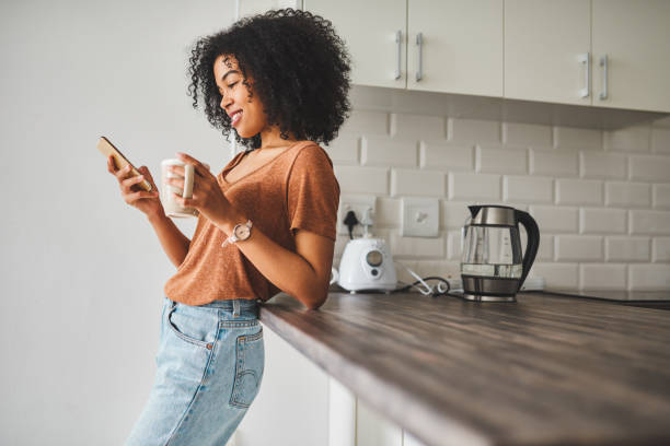 What would the mornings be without social media? Shot of a young woman using a smartphone and having coffee in the kitchen at home curley cup stock pictures, royalty-free photos & images