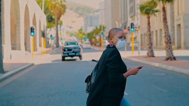 Whatever happens in the world, business will go on Shot of a young businesswoman wearing a mask and using a smartphone while crossing a city street south africa covid stock pictures, royalty-free photos & images