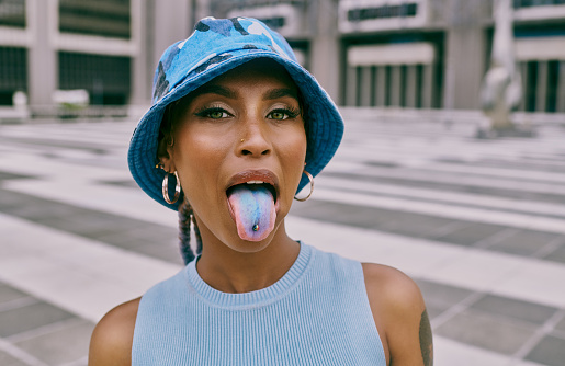 Shot of a trendy young woman sticking out her blue coloured tongue and showing her piercing against an urban background