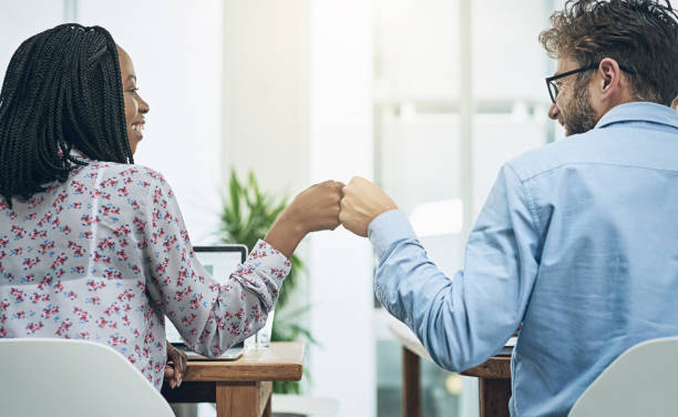 When coworkers become friends Shot of two young colleagues giving each other a fist bump at their desks in a modern office good news stock pictures, royalty-free photos & images