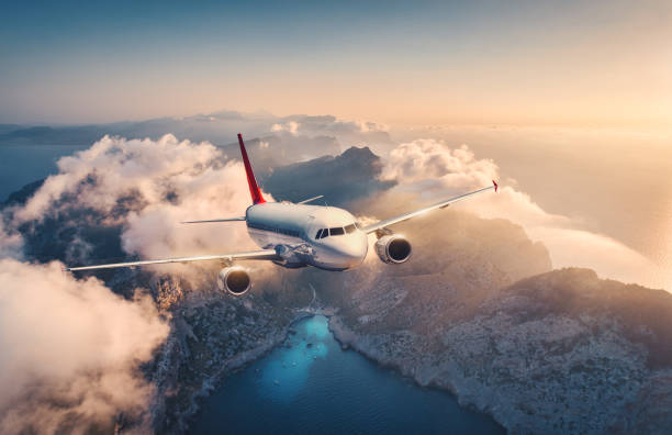 Photo of White airplane is flying over mountains and low clouds at sunset in summer. Landscape with beautiful passenger airplane, sky in clouds, sea, sunlight. Travel. Commercial plane. Aerial view of aircraft