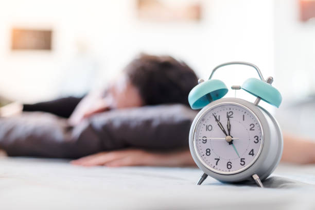 White alarm clock in the morning. Young man sleeps in the background. stock photo