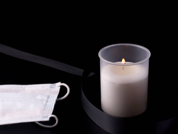 white burning candle next to a black mourning ribbon and a protective medical mask on a black background. concept of death from a pandemic, virus and disease. - covid cemiterio imagens e fotografias de stock