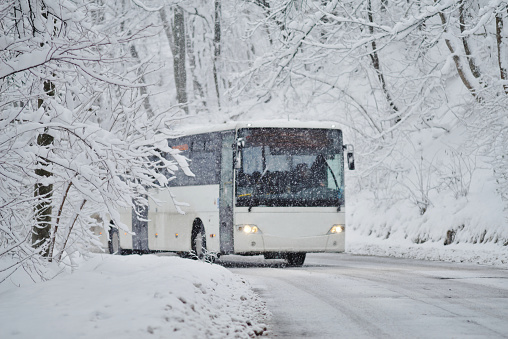 White coach bus on a mountain road through a forest in extreme winter weather.