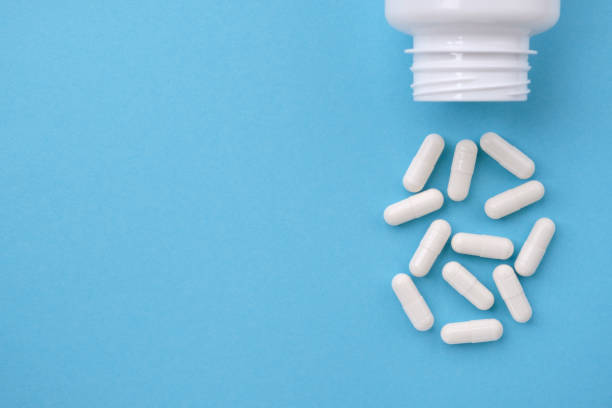 capsule bianche che cadono dalla bottiglia bianca su sfondo di carta blu. concetto di farmacia - farmaco foto e immagini stock
