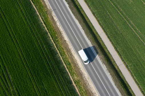 Aerial view of a white delivery van speeding on the road between green fields in spring.