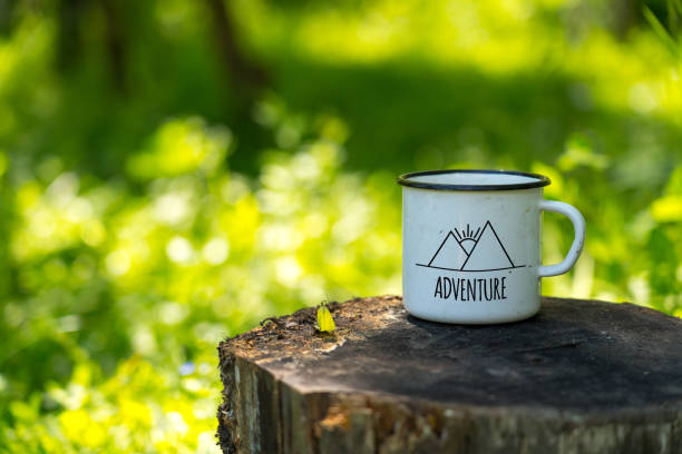 white enamel cup with mountains drawing and adventure text on aged stump in wild forest stock photo