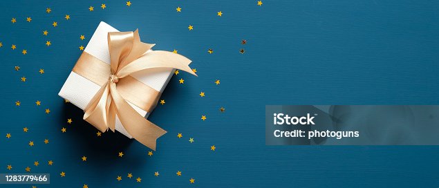 istock White gift box with golden ribbon bow on blue background with confetti stars. Christmas present, valentine day surprise, birthday concept. Flat lay, top view, copy space. 1283779466