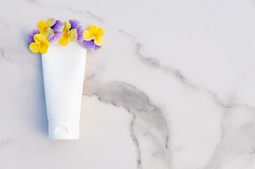 White squeeze bottle cosmetic cream tube and pansy flowers on marble background with copy space. Top view. Unbranded lotion, balsam, hand creme, moisturizer mockup