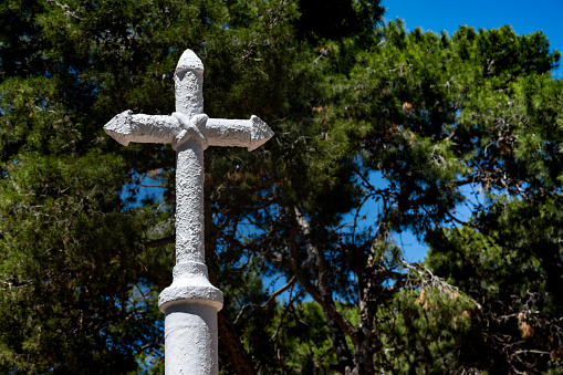 White painted stone christian cross with pine trees in the background. Horizontal