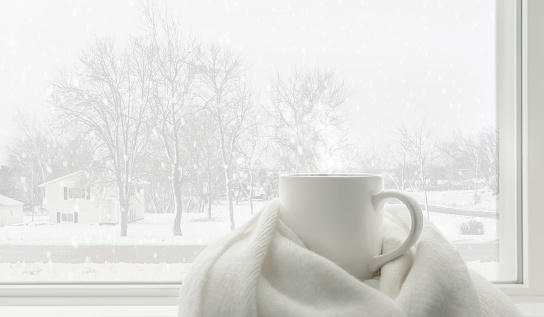 Hot tea mug covered by white comfort soft wool scarf in front of frosted glass window in warm and cozy living room with lonely but beautiful landscape view snow falling outside. White winter concept.