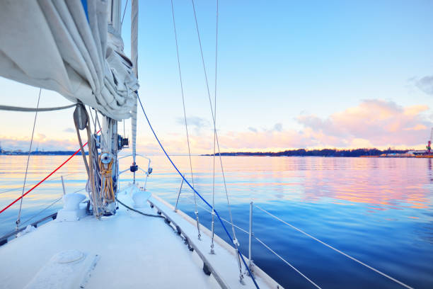 White yacht sailing in a still water at sunset. Frost and first snow on the deck, close-up view to the bow, mast, ropes and sails. Clear blue sky with colorful winter clouds. Norway stock photo