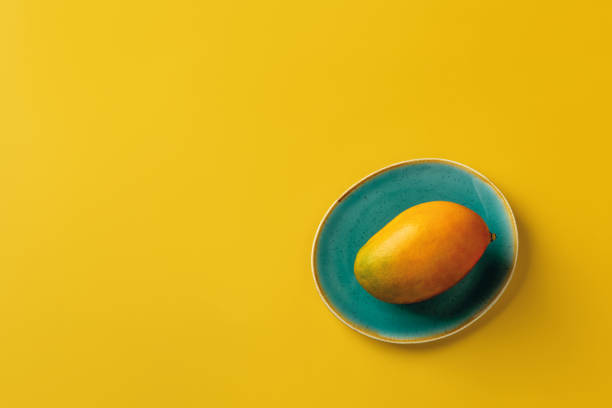 Whole ripe yellow Palmer mango on the turquoise plate over bright yellow background. Ready to eat sweet delicious summer fruit of Mangifera Indica flat lay. Copy space. stock photo