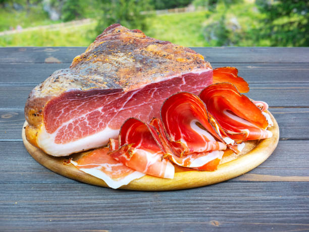 Whole speck with slices. Typical South Tyrolean raw ham outdoors. Italian food. Table in nature, in the woods. stock photo