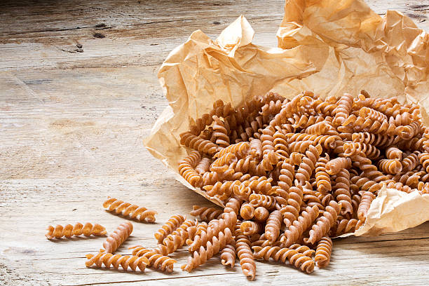 wholemeal pasta fusilli from organic whole grain spelt on paper wholemeal pasta fusilli from organic whole grain spelt falling from a paper bag on a rustic wooden table, copy space wholegrain stock pictures, royalty-free photos & images