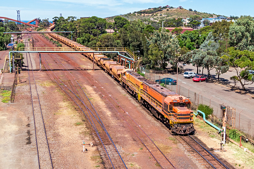 Aerial view of empty iron ore train waiting to depart steel mill unloading yard at Whyalla and travel to one of the mines to load more ore. In the background the line of iron ore wagons recedes back to the unloader. Red dust covers the ground around the railway tracks. ID & logos edited