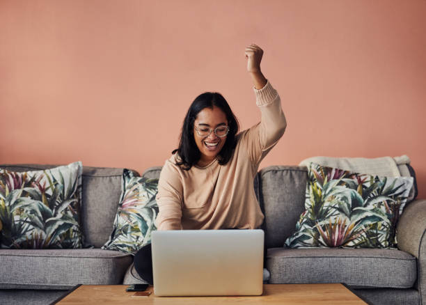 A win might be just what you need Shot of a young woman cheering while using a laptop on the sofa at home good news stock pictures, royalty-free photos & images