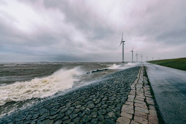Wind turbines on land and offshore in a storm with waves hitting a levee stock photo