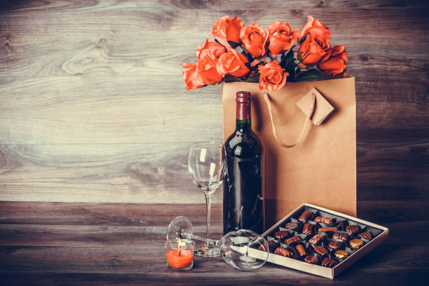 Wine and box of chocolates Red wine bottle, box of chocolates, roses in a paper bag on wooden table. Valentines day celebration concept. Copy space. happy birthday wine bottle stock pictures, royalty-free photos & images