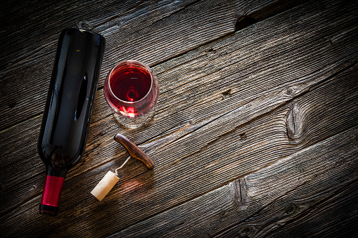 Top view of a wine bottle, wineglass with a corkscrew and a cork stopper shot on rustic textured wooden table. The wineglass is at the left of the table the table and the wine bottle is laying down beside  it. Copy space available for text and/or logo. Predominant color is brown. Low key DSRL studio photo taken with Canon EOS 5D Mk II and Canon EF 100mm f/2.8L Macro IS USM.