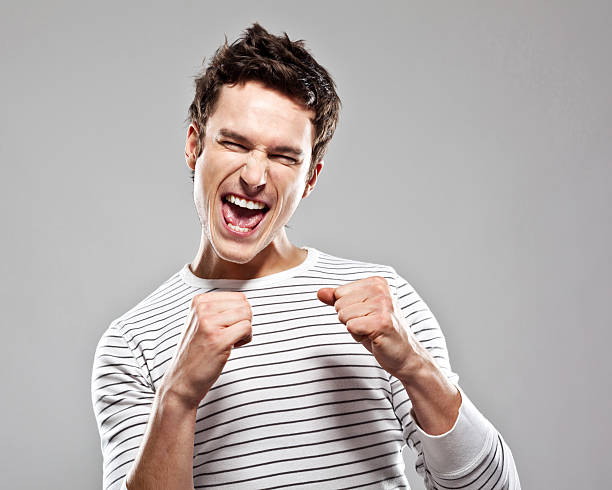 "Portrait of excited young man, raising his fists and screaming with eyes closed. Studio shot, grey background."