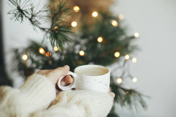 Winter hygge. Hands in cozy sweater holding coffee cup at warm lights. Merry Christmas! stock photo