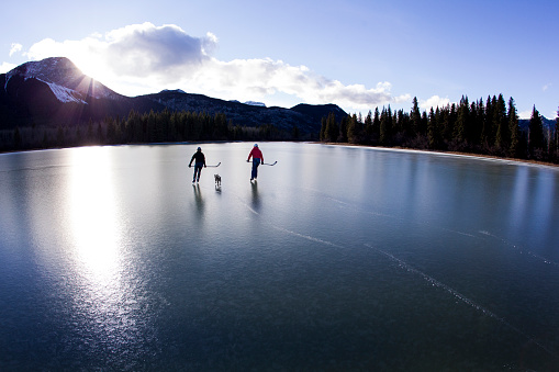 Two women enjoy a winter skate on an outdoor pond in the Rocky Mountains of Canada. They are both carrying hockey sticks and are accompanied by their pet dog.