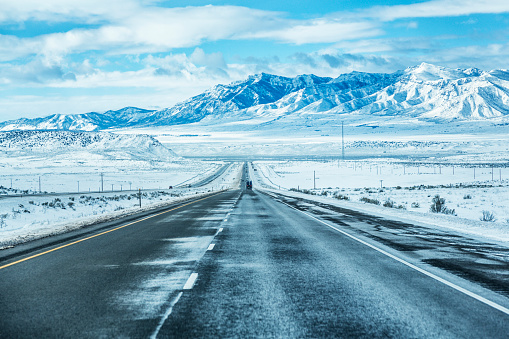 Long, long diminishing perspective downhill straightaway on the southbound Interstate Route 15 expressway highway in southern Utah, USA in early February. Crosswinds are blowing snow sideways, making the asphalt frozen and slippery. The road eventually disappears into a winter snow-blanketed flat valley plain in the distance; with gradually rising, but ultimately very high mountain range peaks in the far background.