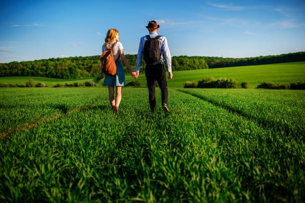 With a backpack, a man in a hat and a woman with long hair go along the path. A couple walks along the meadow stock photo
