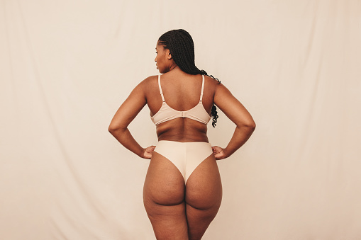 Woman adjusting her underwear in a studio. Rearview of a young woman embracing her natural body and curves. Self-confident young woman wearing beige underwear against a studio background.
