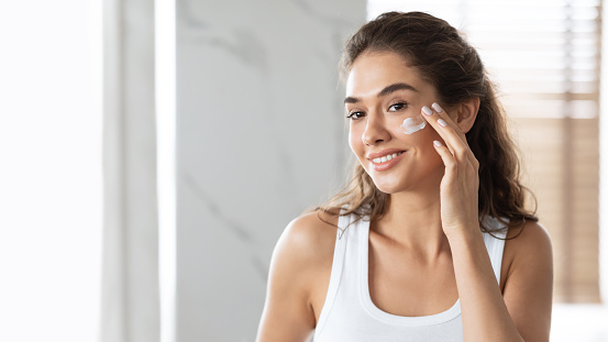 Facial Skincare And Beauty. Woman Applying Face Cream Moisturizer Caring For Skin Standing In Bathroom Indoors, Smiling To Camera. Panorama, Empty Space For Text
