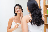 Woman caring of her beautiful skin on the face standing near mirror in the bathroom. Beautiful young woman applying moisturizer on her face. Smiling girl holding little jar of skin cream and applying lotion on face.