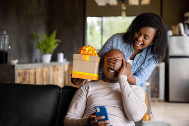 Woman at home surprising her husband with a gift African American woman at home surprising her husband with a gift and covering his eyes fathers day stock pictures, royalty-free photos & images